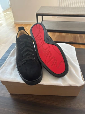 Svarta sneakers från Christian Louboutin - Snygga svarta sneakers i mocka från Christian Louboutin med klassisk röd sula och svarta skosnören. Låg modell med rund tå och diskreta sömmar. Insidan är fodrad i beige läder för extra komfort. Perfekta för dig som vill ha en clean och exklusiv look.