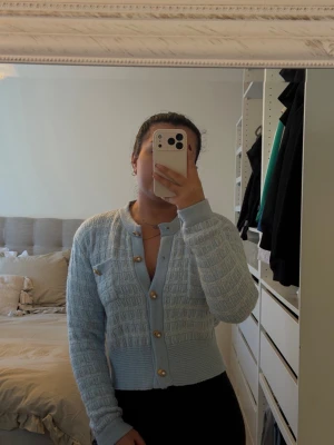 Ljusblå kofta med gulddetaljer - Trendig ljusblå kofta med dekorativa guldfärgade knappar och diskret rutmönster. Koftan har rund halsringning, långa ärmar och ribbade muddar. Perfekt att styla till både jeans och kjol för en snygg look.