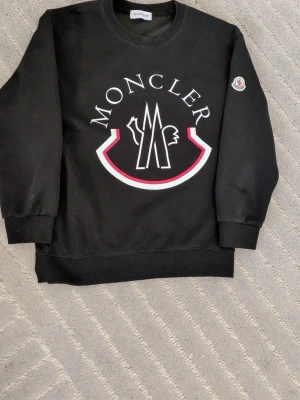 Svart sweatshirt från Moncler - Svart sweatshirt från Moncler med stort vitt och rött logotryck på bröstet och Moncler-märke på ärmen. Tröjan har rund halsringning, ribbade muddar och är tillverkad i mjukt material som känns skönt mot huden. Perfekt för en stilren och trendig look.