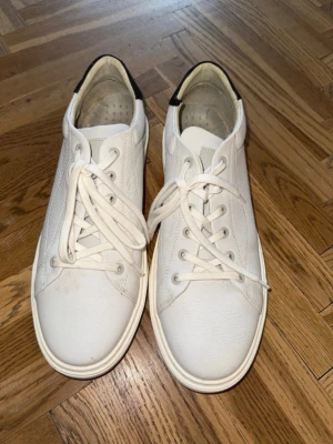 Vita sneakers från G-Star - Snygga vita sneakers från G-Star med klassisk låg profil och svarta detaljer vid hälen. Skorna har vita snören, diskret logga bak och är tillverkade i slittåligt läder med struktur. Perfekta för en clean och stilren look. Liten defekt, se sista bilden.