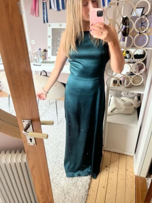 Mörkgrön långklänning med slits - Säljer en elegant mörkgrön långklänning i satinliknande material. Klänningen är ärmlös, har en hög halsringning och en snygg slits på sidan. Perfekt för dig som vill ha en stilren och trendig look.