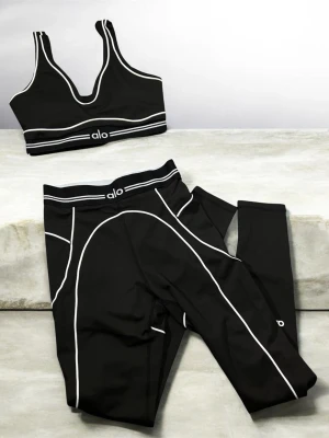Svart träningsset från Alo Yoga - Snyggt svart träningsset från Alo Yoga med vita kontrastsömmar. Setet består av en sport-bh med breda axelband och ett par tights med hög midja. Båda plaggen har Alo-loggan framtill och är tillverkade i stretchigt funktionsmaterial som passar perfekt till gymmet eller yoga.