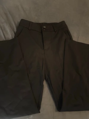 Svarta vida kostymbyxor - Säljer ett par svarta kostymbyxor med hög midja och vida ben. Byxorna har resår bak i midjan, bälteshällor och knappstängning fram. Perfekta för en clean och stilren look. Materialet känns mjukt och följsamt.