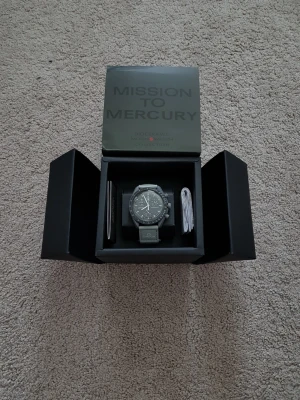 Omega x Swatch MoonSwatch Mercury - Säljer en Omega x Swatch MoonSwatch Mission to Mercury. Klockan har en mörkgrå boett och svart urtavla med vita detaljer. Bandet är grått med Omega-logga. Kommer i originalbox med tillbehör. Perfekt för dig som gillar ikoniska samarbeten.