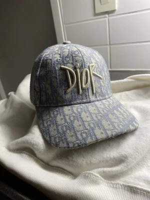 Blå Dior keps med monogram - Säljer en snygg blå keps från Dior med klassiskt monogrammönster och stor broderad Dior-logga framtill. Kepsen är i denimliknande material och har justerbar rem bak för perfekt passform. Perfekt accessoar för dig som gillar streetstyle och lyx.