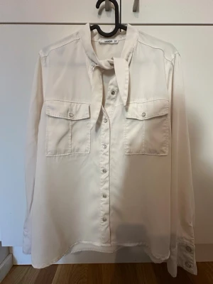 Vit blus med knytband från Mango M - Stilren vit blus från Mango med knytband i halsen och två bröstfickor med lock. Blusen har knappar framtill och långa ärmar med manschett och tryckknappar. Perfekt till jeans eller kjol för en trendig look.