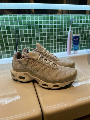 Nike Air Max Plus beige sneakers - Säljer ett par Nike Air Max Plus sneakers i beige med mesh- och syntetmaterial. Skorna har ikoniska vågiga detaljer, synlig Air-dämpning i sulan och vita snören. Perfekta för dig som gillar streetwear och vill ha en clean look.