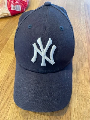 Mörkblå New Era NY Yankees keps - Snygg mörkblå keps från New Era med klassisk New York Yankees-logga broderad i vitt framtill. Modellen är 9FORTY med böjd skärm och justerbar rem bak. Tillverkad i slitstarkt bomullsmaterial, perfekt för en sportig och avslappnad stil.