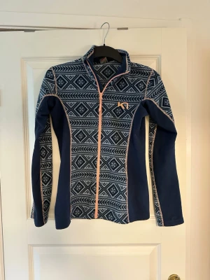 Blå mönstrad fleecejacka Kari Traa Stl xs  - Snygg blå fleecejacka från Kari Traa med hel dragkedja och grafiskt mönster i vitt. Jackan har rosa detaljer längs dragkedjan och sömmarna samt hög krage. Perfekt för lager-på-lager och outdoor-aktiviteter.