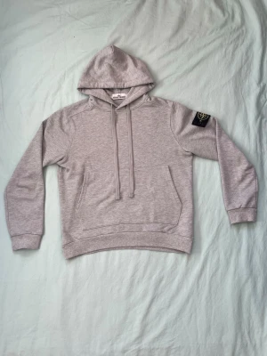 Grå hoodie från Stone Island - Snygg grå hoodie från Stone Island med klassisk huva och dragsnören. Har en stor ficka framtill och den ikoniska Stone Island-loggan på vänster ärm. Tillverkad i mjukt bomullsmaterial som känns skönt mot huden. Perfekt för en avslappnad och trendig stil.