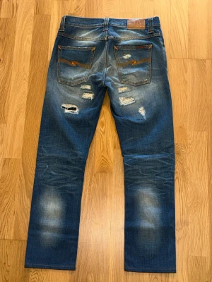  Nudie Jeans Grim Tim - Säljer ett par blå jeans från Nudie Jeans med fades och trasiga detaljer både fram och bak. Jeansen har raka ben, klassisk femficksmodell och kontrastsömmar på bakfickorna. Materialet är denim i bomull och de har en cool, använd look. Kontaka mig vid fundering av pris eller storlek!