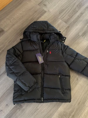 Svart pufferjacka från Polo Ralph Lauren - Svart pufferjacka från Polo Ralph Lauren med huva och dragkedja framtill. Jackan har två sidofickor med dragkedja och den klassiska röda logotypen broderad på bröstet. Tillverkad i ett glansigt, vadderat material som håller värmen under kalla dagar.