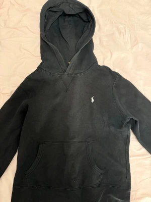 Svart hoodie från Polo Ralph Lauren - Klassisk svart hoodie från Polo Ralph Lauren med huva och känguruficka. Broderad logga på bröstet i vitt. 