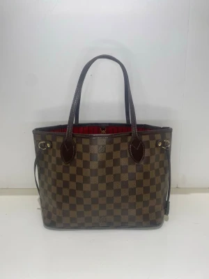 Louis Vuitton Neverfull Damier Ebene PM handväska - Snygg handväska från Louis Vuitton i klassiskt brunt Damier Ebene-mönster. Väskan har två långa handtag, detaljer i mörkbrunt läder och rödrandigt innerfoder. Praktisk öppen modell med sidoremmar och guldfärgade metalldetaljer.