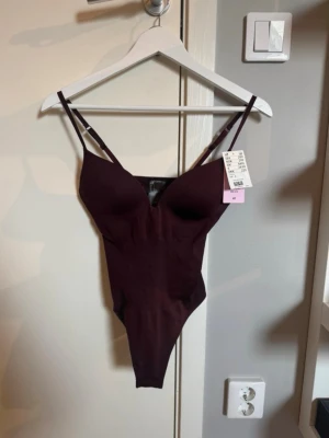Mörklila body med smala axelband - Snygg mörklila body med smala justerbara axelband och vadderade kupor. Tillverkad i ett mjukt, stretchigt material som sitter tight mot kroppen. Perfekt för att skapa en clean och stilren look under kläder.