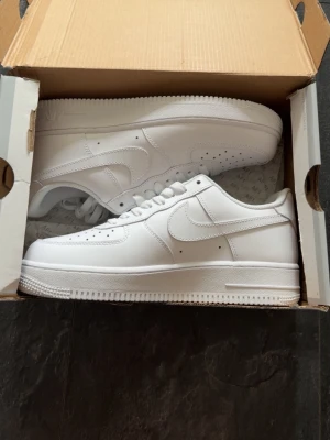 Nike Air Force 1 vita sneakers - Klassiska Nike Air Force 1 sneakers i helvitt läder med låg profil och ikonisk swoosh på sidan. Skorna har perforerade detaljer på tån för ventilation och en tjock, räfflad sula för extra komfort. Perfekta för dig som gillar stilrena och tidlösa sneakers.