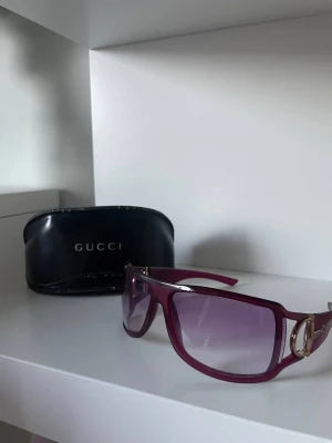 Lila solglasögon från Gucci - Snygga lila solglasögon från Gucci med bred båge och guldfärgade detaljer på sidorna. Glasen är tonade i lila och bågarna har en cool, modern form. Kommer med originalfodral från Gucci.