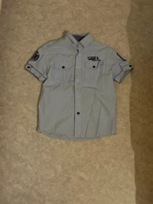 Ljusblå skjorta med patch och knappar - Kortärmad ljusblå skjorta från Sport 4 med knappar framtill, två bröstfickor och coola tygpatchar på ärmarna. Skjortan har klassisk krage och detaljer som ger en avslappnad men ändå snygg look.