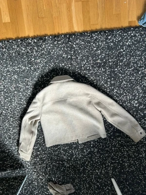 Beige ullkappa från Zara - Kort beige kappa från Zara i ullmix med klassisk krage och raka ärmar. Snygg minimalistisk design och neutral färg som passar till det mesta. Perfekt för dig som gillar stilrena och trendiga ytterplagg.