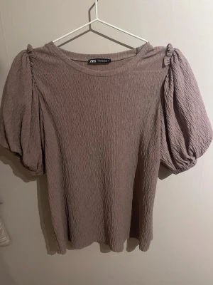 Brun/grå topp med puffärmar från Zara - Trendig ljuslila topp från Zara med puffiga korta ärmar och rund halsringning. Tillverkad i ett krinklat, stretchigt material. Knappt använd!!💕