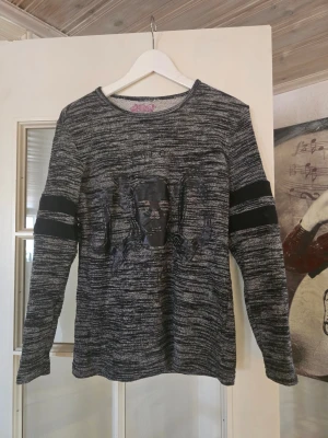 Gråmelerad tröja extreme clothing  - Cool långärmad tröja i gråmelerat tyg med svart döskalletryck framtill och svarta detaljer på ärmarna. Rund halsringning och avslappnad passform. Perfekt för dig som gillar edgy streetstyle.