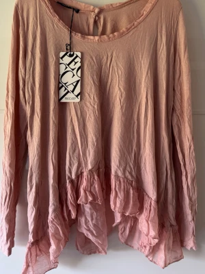 Ny. Rosa volangblus från Peccati - Supersöt rosa blus från Peccati med volangdetaljer och asymmetrisk nederkant. Blusen har lång ärm och öppen rygg med knappstängning upptill. Perfekt för dig som gillar en romantisk och bohemisk stil. Storlek xs, men stort i storlek och kan passa s/m också.