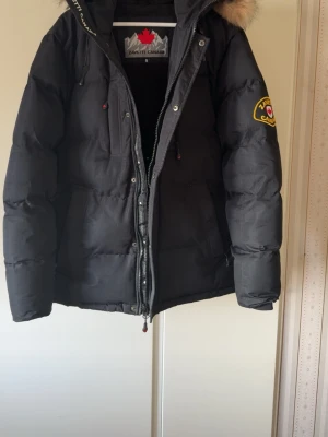 Svart dunjacka från Zavetti Canada - Snygg svart dunjacka från Zavetti Canada med patch på ärmen och avtagbar huva med fuskpäls. Jackan har dragkedja och tryckknappar framtill samt flera fickor. Perfekt för kalla vinterdagar och riktigt varm.