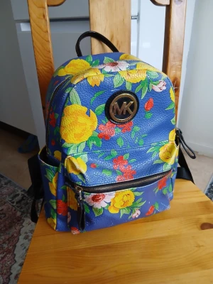 Blommig ryggsäck  - Färgstark ryggsäck i blått konstläder med stora gula, röda och vita blommor. Har ett stort fack, framficka med dragkedja och sidofickor. 