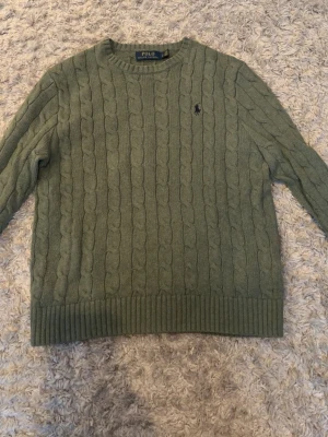 Olivgrön kabelstickad tröja Polo Ralph Lauren - Snygg olivgrön kabelstickad tröja från Polo Ralph Lauren med rund halsringning och klassisk broderad logga på bröstet. Tröjan har ribbade muddar vid ärmslut och nederkant, perfekt för dig som gillar tidlös och stilren design.