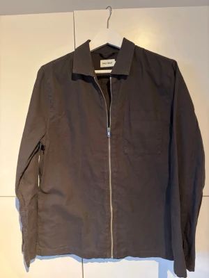 Mörkbrun overshirt från East West - Stilren mörkbrun overshirt från East West med dragkedja framtill och en bröstficka. Overshirten har klassisk krage och raka ärmar. Tillverkad i ett mjukt bomullstyg som ger en avslappnad look.