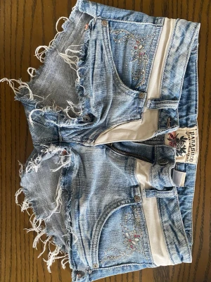 Ljusblå lågmidjade jeansshorts - Säljer ett par ljusblå jeansshorts från Parasuco med råa fransiga kanter, broderade detaljer och dekorativa fickor med glitterdetaljer💐🥂. Shortsen har en mycket låg midja  och klassisk femficksdesign🤩🤩🤗🤗. Perfekta till sommaren, våren miiih💓🫶🙌Storlek 27 och passar strl 34-36💃