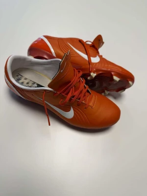 Nike mercurial vapor 1 Remake - Säljer ett par snygga orange fotbollsskor från Nike med vita detaljer och klassisk swoosh på sidan. Skorna har snörning, låg profil och fasta dobbar för bra grepp på planen. Ovandelen är i syntetmaterial och insidan är vit. Perfekta för dig som vill sticka ut på fotbollsplanen!