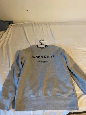 Grå sweatshirt från Björn Borg - Snygg grå sweatshirt från Björn Borg med tryck på bröstet där det står 'BJÖRN BORG STOCKHOLM 1956'. Tröjan har rund halsringning och långa ärmar. Tillverkad i mjukt material som passar perfekt till en avslappnad stil.