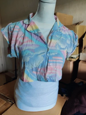 Pastellfärgad regnbåge 🌈 kort blus med mönster - Kort blus i tunnt tyg med pastellfärgade moln och regnbågsmönster. Skjortkrage och knäppning framtill. Blusen har korta ärmar och avslappnad passform, perfekt för en färgglad och lekfull stil.