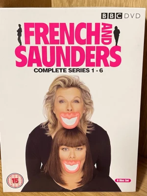 French and Saunders Complete Series  DVD-box - French and Saunders Complete Series 1-6 DVD-box med 6 skivor. Omslaget är vitt med stora rosa bokstäver och bilder på Dawn French och Jennifer Saunders. Perfekt för dig som gillar brittisk humor och klassiska sketcher.