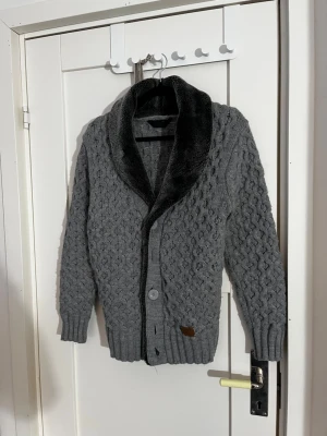 Grå stickad kofta med sjalkrage - Snygg grå stickad kofta med grovt mönster och bred, mjuk sjalkrage i mörkare material. Koftan har knäppning framtill med stora knappar och ribbade muddar vid ärmslut och nederkant. Perfekt för lager på lager och chill stil.