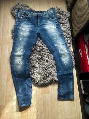 Blå slitna jeans från G-Star RAW - Säljer ett par blå jeans från G-Star RAW med slitningar och distressed detaljer. Jeansen har klassisk femficksmodell, dragkedjegylf och smal passform. Perfekta för en avslappnad och trendig look.