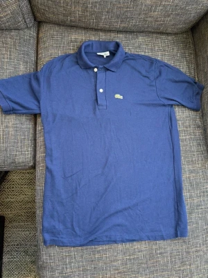 Marinblå pikétröja från Lacoste - Klassisk marinblå pikétröja från Lacoste med två vita knappar och den ikoniska gröna krokodilloggan på bröstet. Tröjan är gjord i bomull och har korta ärmar samt krage. Perfekt för en clean och sportig stil.