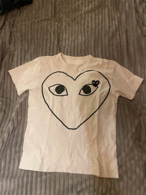 Vit Comme des Garçons Play t-shirt - Säljer en vit t-shirt från Comme des Garçons Play med det ikoniska hjärtmotivet och ögon på bröstet. T-shirten har rund hals och korta ärmar. Materialet är mjuk bomull och trycket är i svart med en broderad detalj. Har blivit lagad vid nacken. Storlek S med passar XS. 
