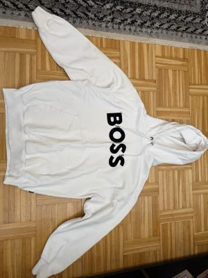Vit hoodie från BOSS med tryck - Säljer en clean vit hoodie från BOSS med stor svart logga på bröstet. Hoodien har huva med dragsko och en klassisk magficka. Perfekt för dig som gillar stilrena plagg med tydlig branding.
