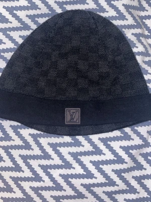  Louis Vuitton mössa - Snygg mörkgrå mössa från Louis Vuitton med diskret rutmönster och LV-logotyp på en patch framtill. Mössan är stickad och har en bred ribbad kant som ger en skön passform. Perfekt för dig som vill ha en exklusiv och stilren accessoar. (Äkta)