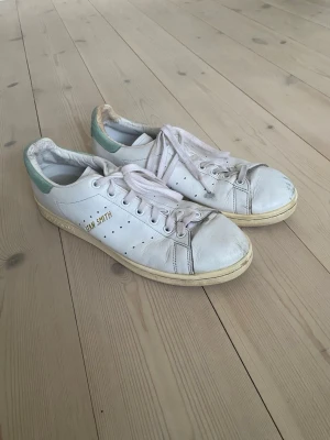 Adidas Stan Smith vita sneakers - Klassiska vita Adidas Stan Smith sneakers i läder med gröna detaljer på hälen och guldfärgad Stan Smith-text på sidan. Skorna har perforerade ränder, vit sula och snörning framtill. Perfekt för en clean och tidlös stil.