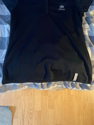 Svart ridtopp från Active Riding Wear - Snygg svart topp från Active Riding Wear med knappar framtill och diskret logga på bröstet. Tillverkad i ett lätt och ventilerande material som passar perfekt för ridning. Klassisk passform och korta ärmar. Passar perfekt som S.