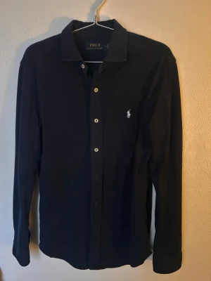 Marinblå skjorta från Polo Ralph Lauren - Klassisk marinblå skjorta från Polo Ralph Lauren med vit broderad logga på bröstet. Skjortan har långa ärmar, knappar framtill och en stilren krage. Perfekt för dig som gillar tidlös och enkel stil.