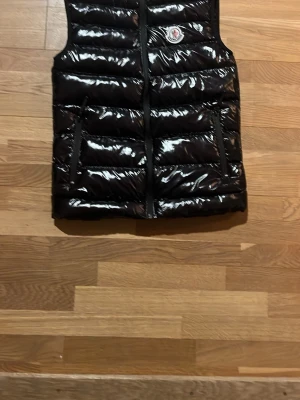 Svart glansig dunväst från Moncler - Snygg svart dunväst från Moncler med glansigt yttermaterial och klassisk logga på bröstet. Västen har två dragkedjeförsedda fickor och quiltad design som ger en modern look. Perfekt att slänga över en hoodie eller tröja för extra värme.