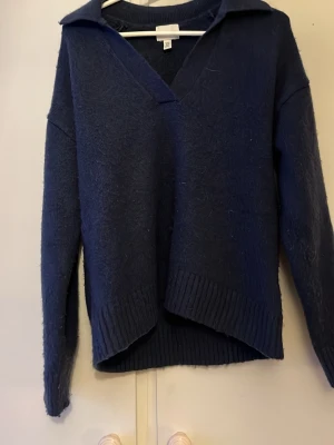 Mörkblå stickad tröja med krage - Mysig mörkblå stickad tröja med v-ringning och bred krage. Tröjan har långa ärmar och ribbade muddar samt en något asymmetrisk nederkant. Perfekt för lager på lager och en avslappnad stil.