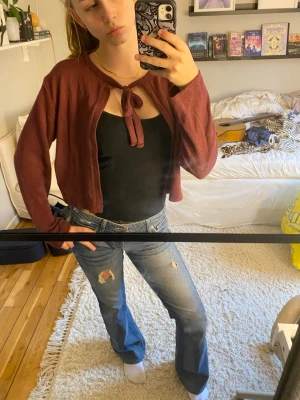 Vinröd knytkofta i croppad modell - Säljer en vinröd, kort kofta med knyt framtill. Koftan har långa ärmar och är gjord i ett mjukt, tunt material som passar perfekt över ett linne eller t-shirt. 