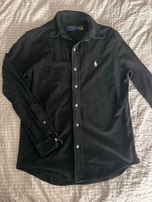 Svart manchesterskjorta Polo Ralph Lauren - Klassisk svart manchesterskjorta från Polo Ralph Lauren med vit broderad logga på bröstet. Skjortan har långa ärmar, knappar framtill och en mjuk, lite tjockare känsla. Perfekt för dig som gillar stilrena plagg med en exklusiv touch.