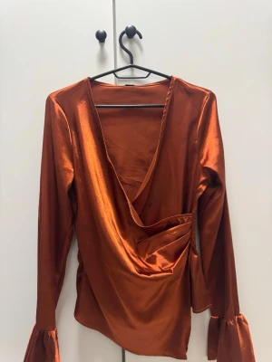 Kopparfärgad omlottblus med volangärm - Snygg kopparfärgad omlottblus i glansigt material med djup v-ringning och långa ärmar med volangdetalj vid ärmslutet. 