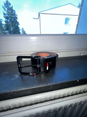 Svart skärp från Tommy Hilfiger - Snyggt svart bälte från Tommy Hilfiger i skinn med klassisk metallspänne. Detaljer i rött, vitt och blått vid spännet som ger en stilren touch. Perfekt accessoar för att lyfta din outfit.